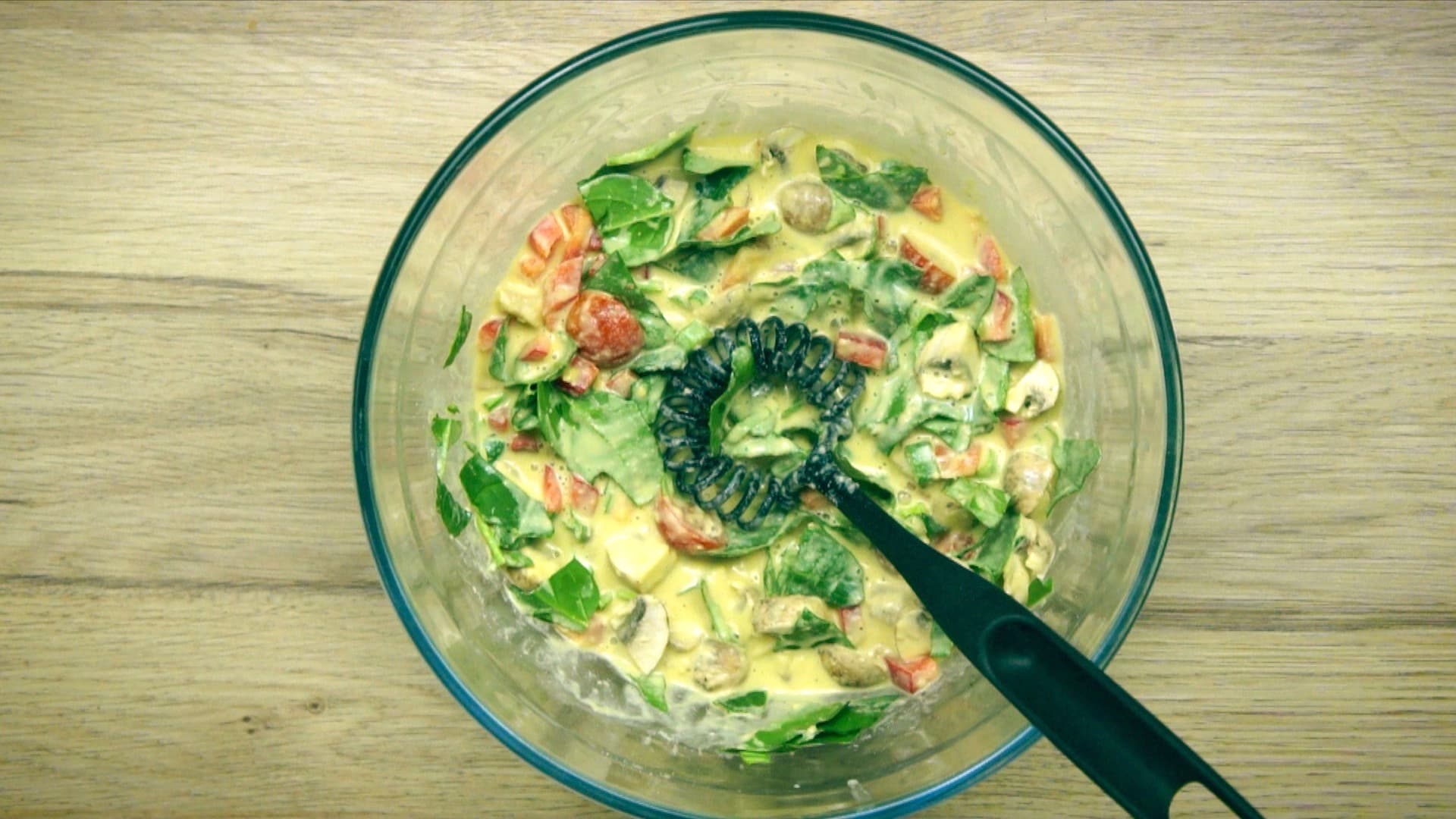 A whisk is placed in a yellow batter full of chopped veggies.