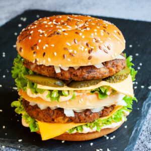 A burger with layers of green ruffled lettuce, yellow sauce, thin cheese slices, dark brown burger patty twice, sliced pickles and white chopped onion.