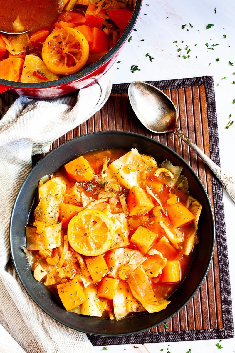 Black plate from above with red tomato-based soup and chopped vegetables like cabbage, orange sweet potato, celery stalk slices, topped with lemon slices and dried green herbs