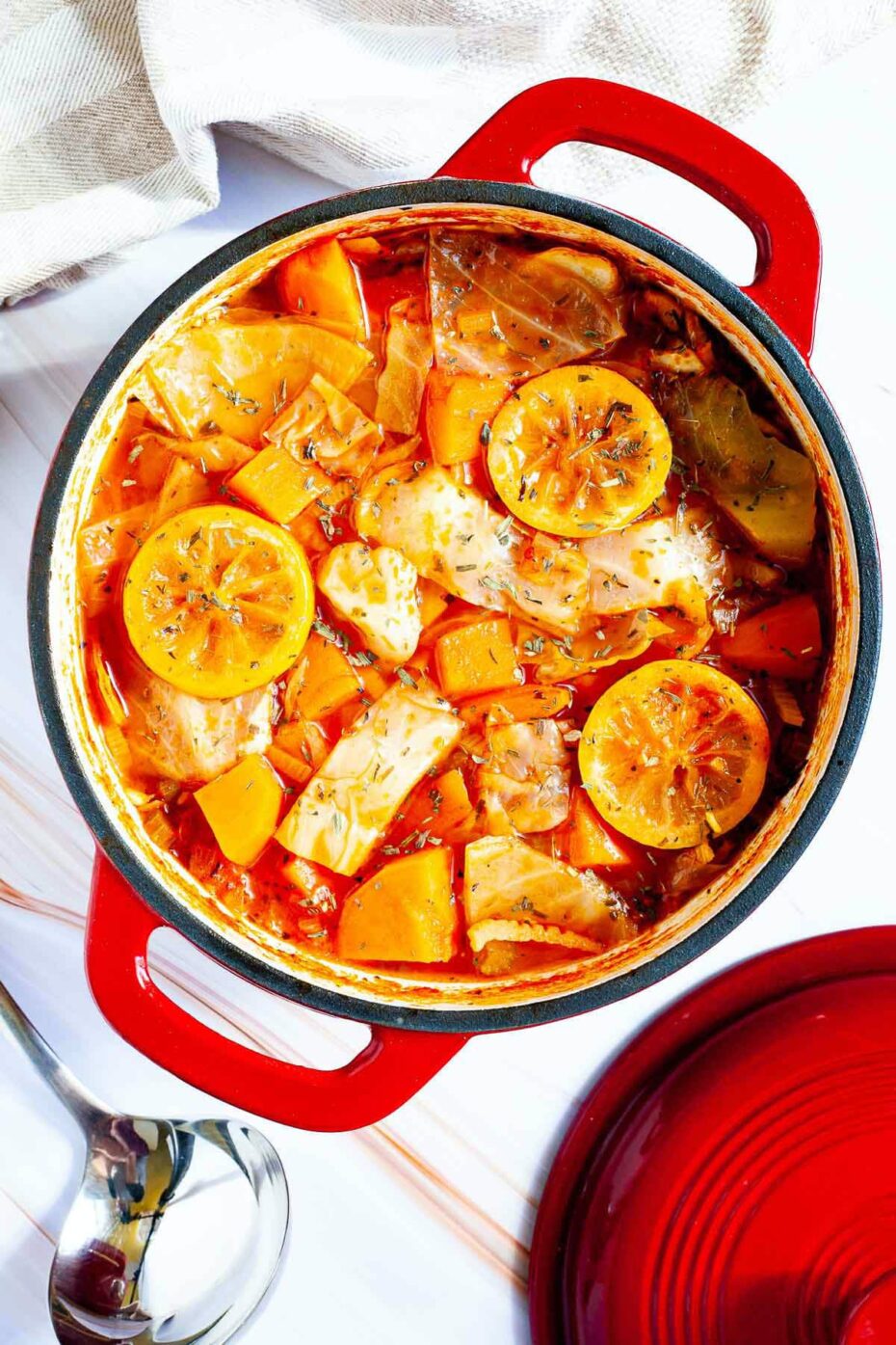 Red dutch oven with red tomato-based soup and chopped vegetables like cabbage, orange sweet potato, celery stalk slices, topped with lemon slices and dried green herbs