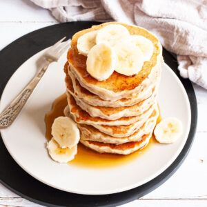White plate with a stack of pancakes topped with banana slices with maple syrup pouring down