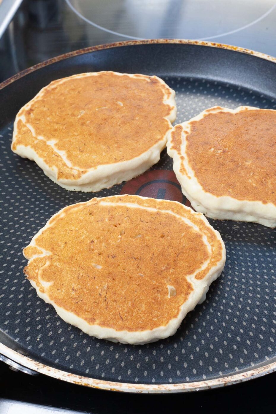 Black frying pan with 3 pancakes after they are flipped so the tops are golden brown
