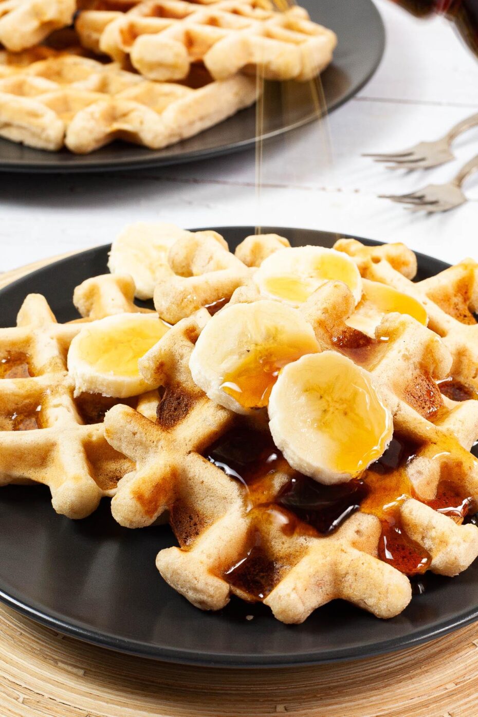 Black plate with a stack of waffles topped with sliced banana and maple syrup is pouring on them. There are 3 waffles on another black plate in the background.