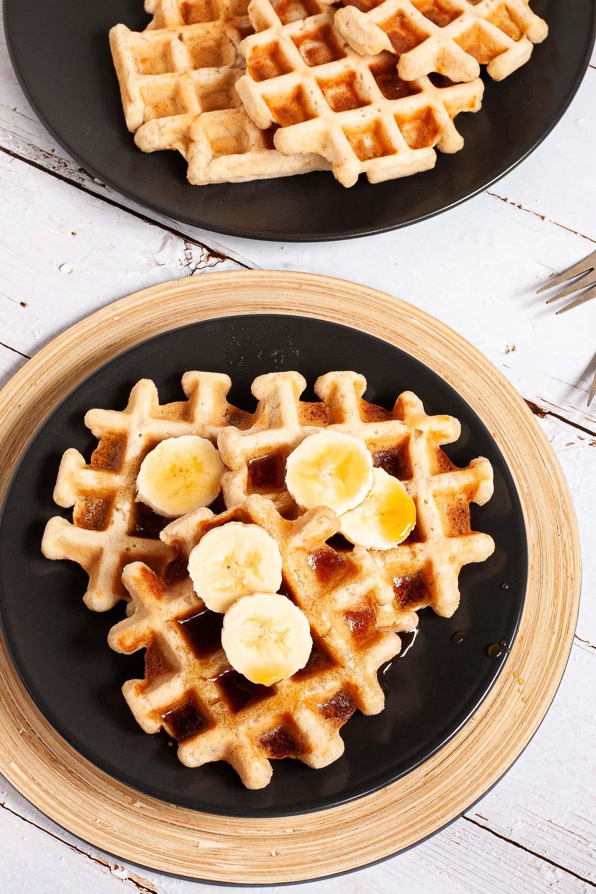 Black plate with a stack of waffles topped with sliced banana and maple syrup from above. There are 3 waffles on another black plate in the background.