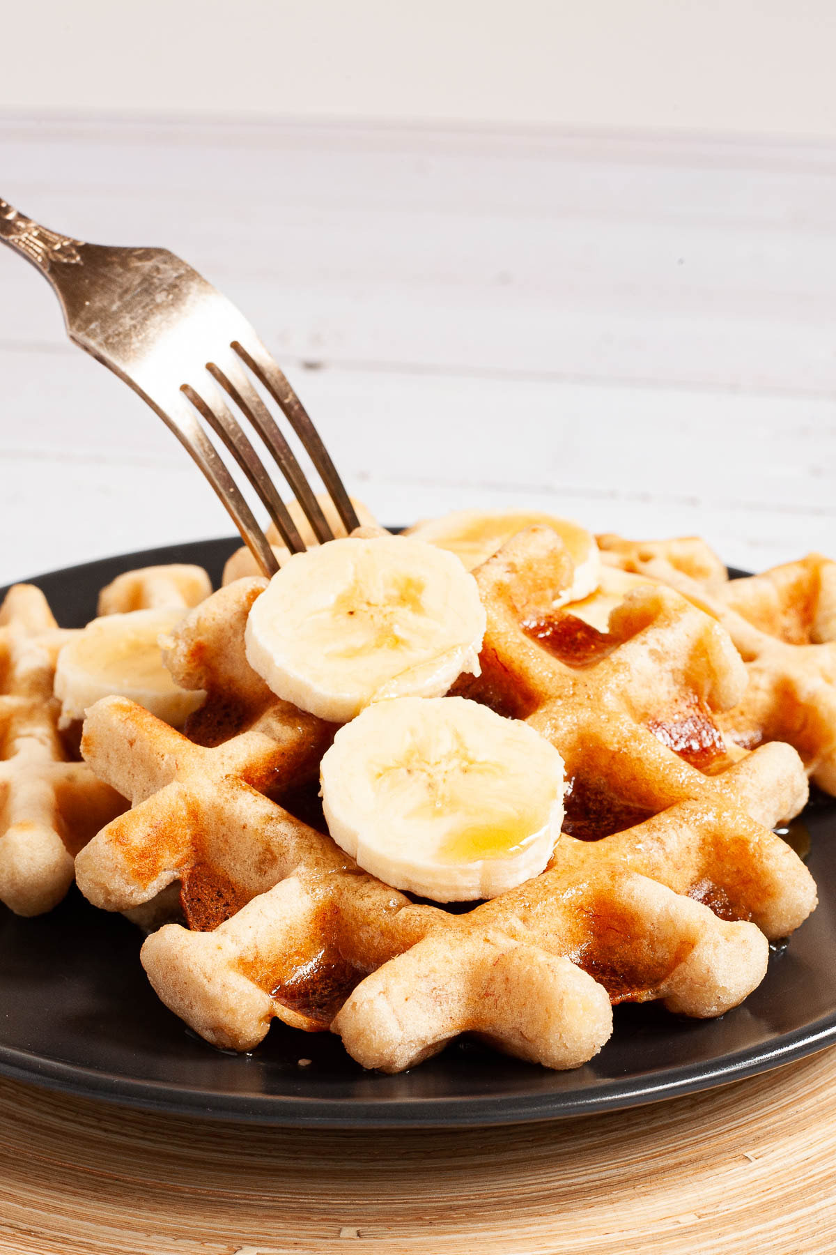 Black plate with a stack of waffles topped with sliced banana and maple syrup. A fork is piecring one in the middle.