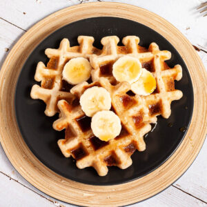 Black plate with a stack of waffles topped with sliced banana and maple syrup pictured from above.
