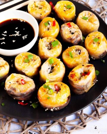 Lots of round-shaped rice paper dumplings arranged in a semi-circular dish facing upwards sprinkled with sesame seeds, chopped spring onion and red hot pepper around a brown dipping sauce.