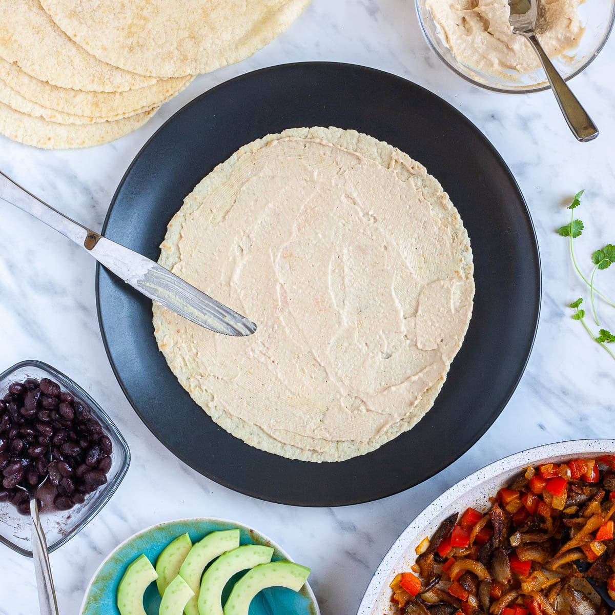 A tortilla covered with light brown hummus on a black plate. The fillings are around it like cooked veggies, avocado slices, black beans, more tortilla and more hummus.