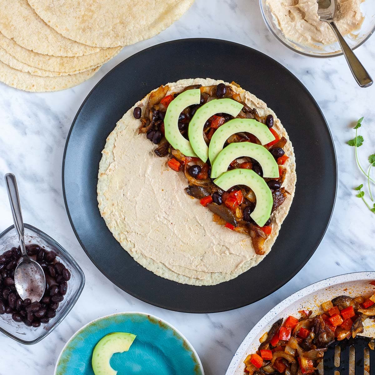 A tortilla covered with light brown hummus, chopped pan-fried veggies, and avocado slices. Leftover fillings are around it with more tortilla and more hummus.