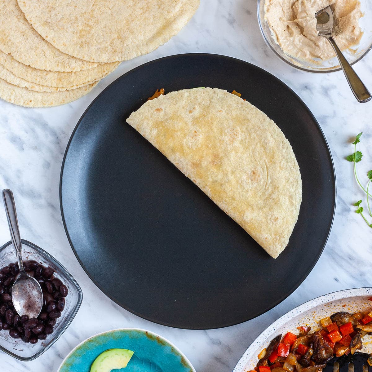 A folded tortilla on a black plate. The fillings are around it like cooked veggies, avocado slices, black beans, more tortilla and more hummus.