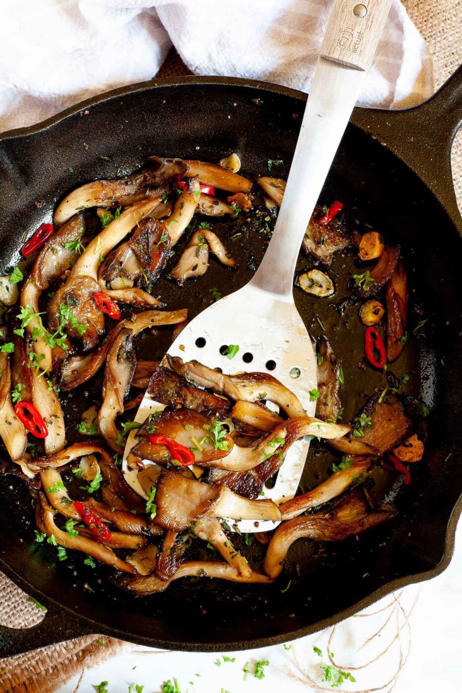 Black cast iron skillet from above with crispy brown oyster mushroom, sliced red pepper, garlic slices and chopped fresh green herbs. A silver slotted turner is taking a serving.