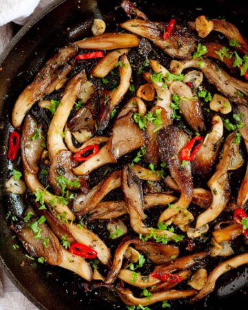 Black cast iron skillet from above with crispy brown oyster mushroom, sliced red pepper, garlic slices and chopped fresh green herbs.