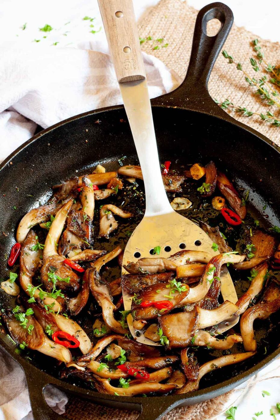 Black cast iron skillet from above with crispy brown oyster mushroom, sliced red pepper, garlic slices and chopped fresh green herbs. A silver slotted turner is taking a serving.