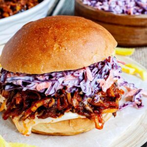 A burger sandwich on a wooden board with white sauce, brown juicy strips and purple cabbage coleslaw. A white bowl is the remaining brown juicy mushroom slices and a wooden bowl with more coleslaw salad and french fries