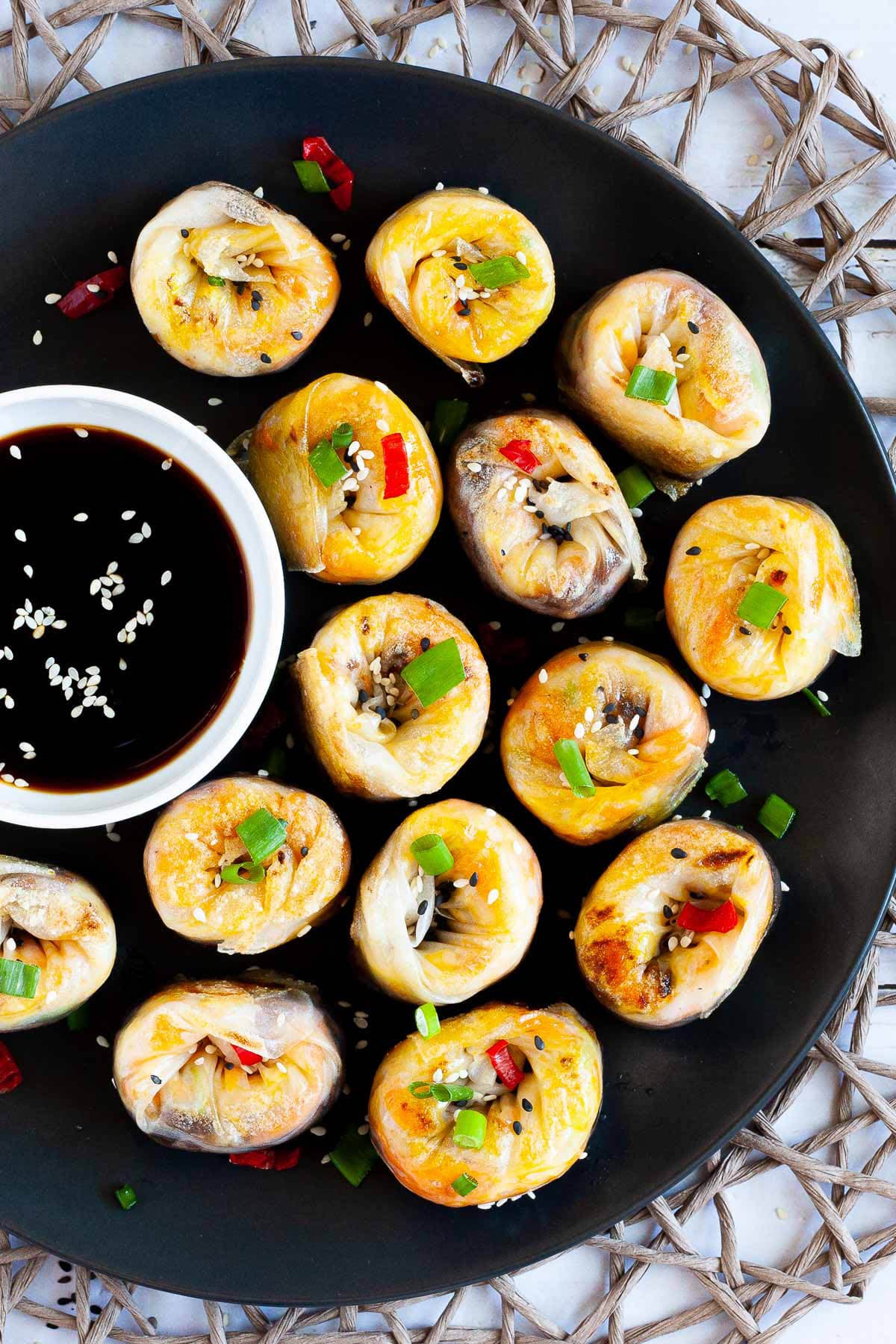 Lots of round-shaped rice paper dumplings arranged in a semi-circular dish facing upwards sprinkled with sesame seeds, chopped spring onion and red hot pepper around a brown dipping sauce.