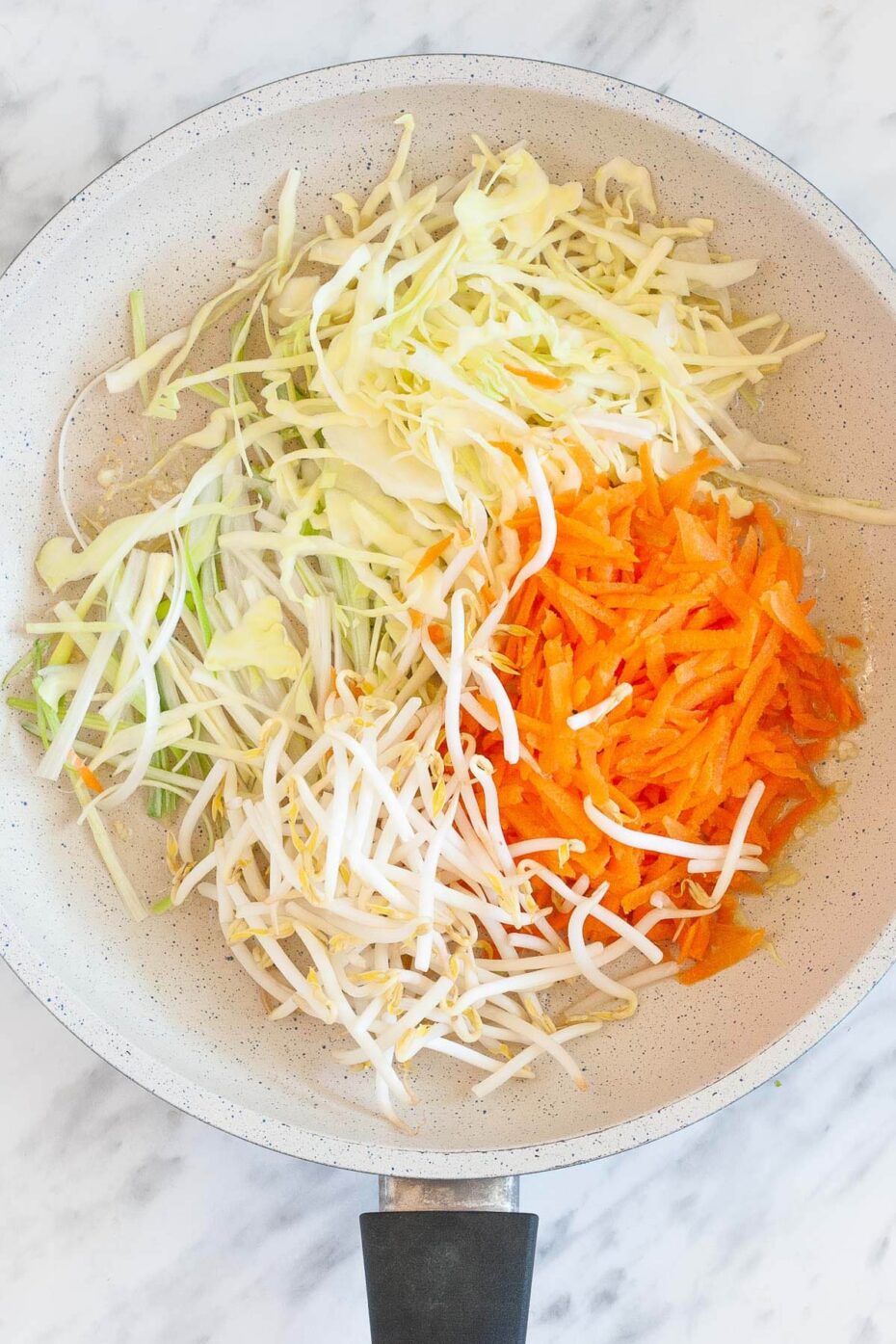 A white frying pan from above full of white and orange shredded veggies like carrots, cabbage, spring onion and sprouts raw before cooking