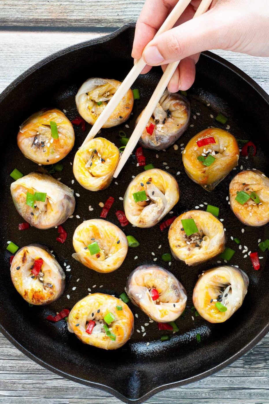 Lots of round-shaped rice paper dumplings in a black cast iron skillet facing upwards sprinkled with sesame seeds, chopped spring onion and red hot pepper around a brown dipping sauce. A hand is holding wooden chopsticks.
