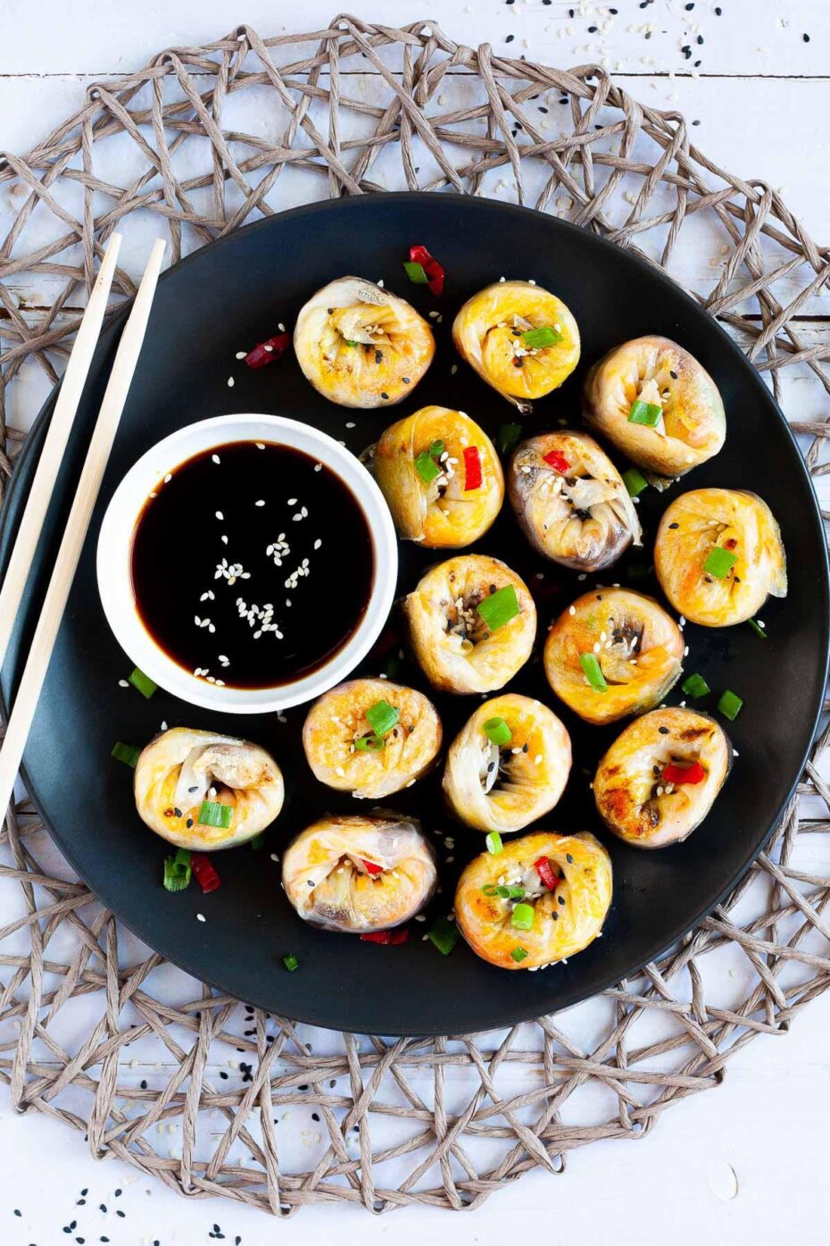 Lots of round-shaped rice paper dumplings arranged in a semi-circular dish facing upwards sprinkled with sesame seeds, chopped spring onion and red hot pepper around a brown dipping sauce. Wooden chopsticks are on the side.