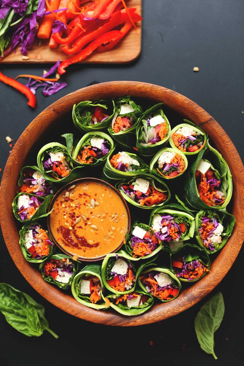 Colorful filling of carrots, tofu, red pepper and purple cabbage are wrapped in dark green leaves and cut in half. They are placed in a round wooden bowl with a small bowl of peanut sauce. 