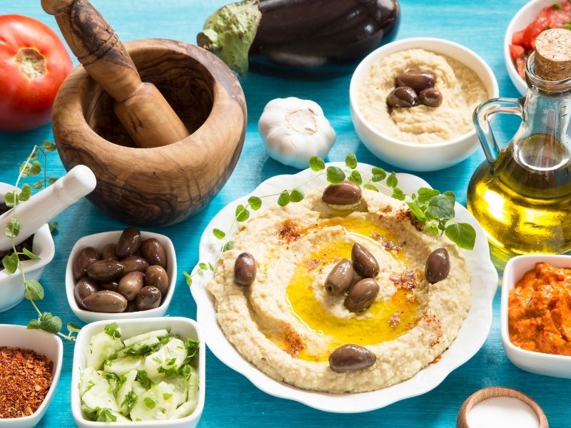 Hummus with olives on a white plate and many small white bowls are around with ingredients and spices. 