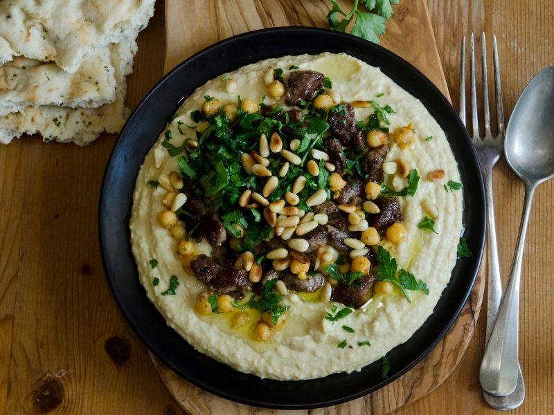 Hummus on a black plate with toasted pine nuts and roasted mushrooms, chickpeas and freshly chopped parsley
