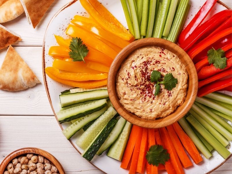 Hummus in a small brown bowl in the middle of a white plate with vegetables sticks all around it