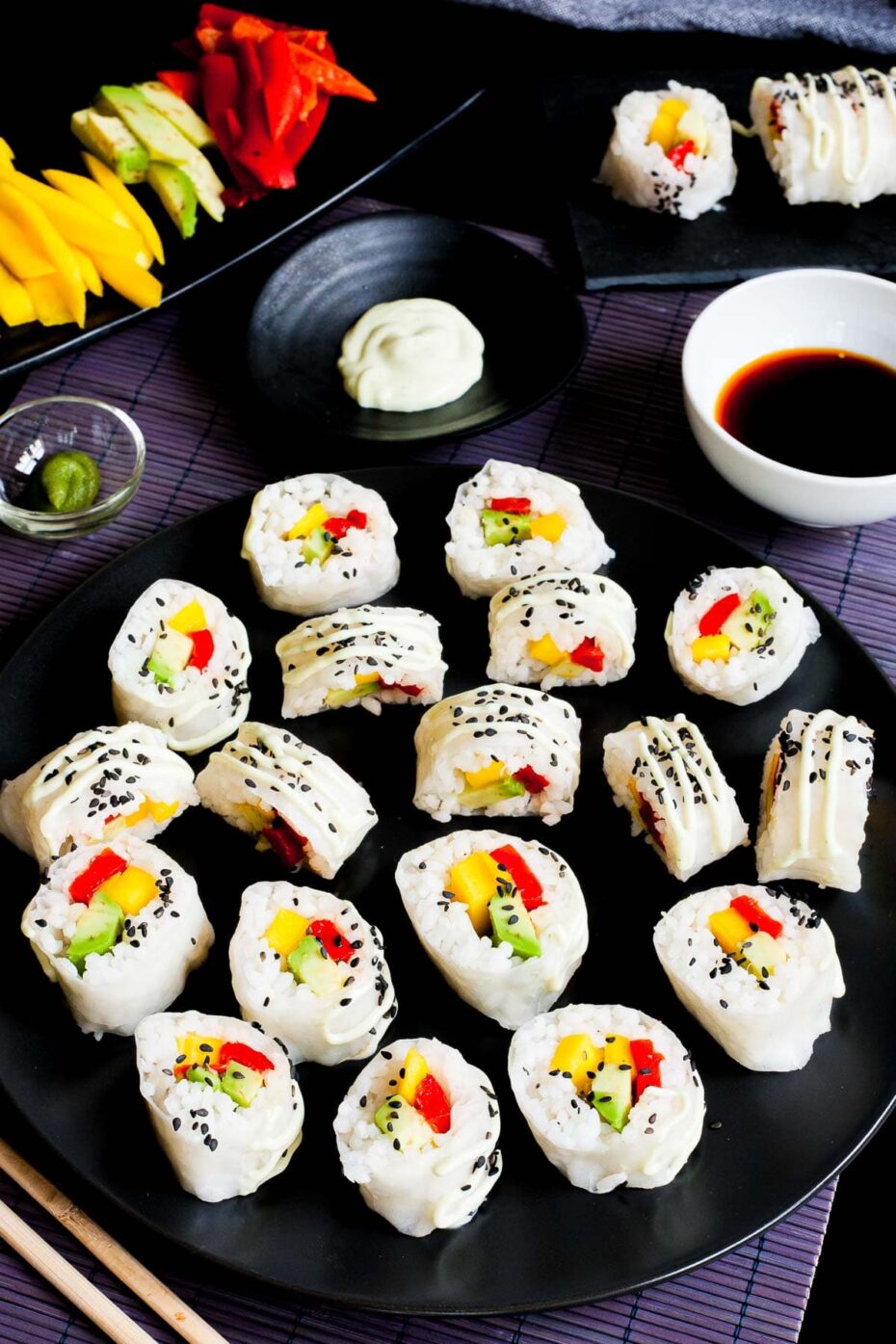 Large black round plate with a lot of white sushi rolls with colorful filling sprinkled with tiny black sesame seeds. Condiment bowls are around it.