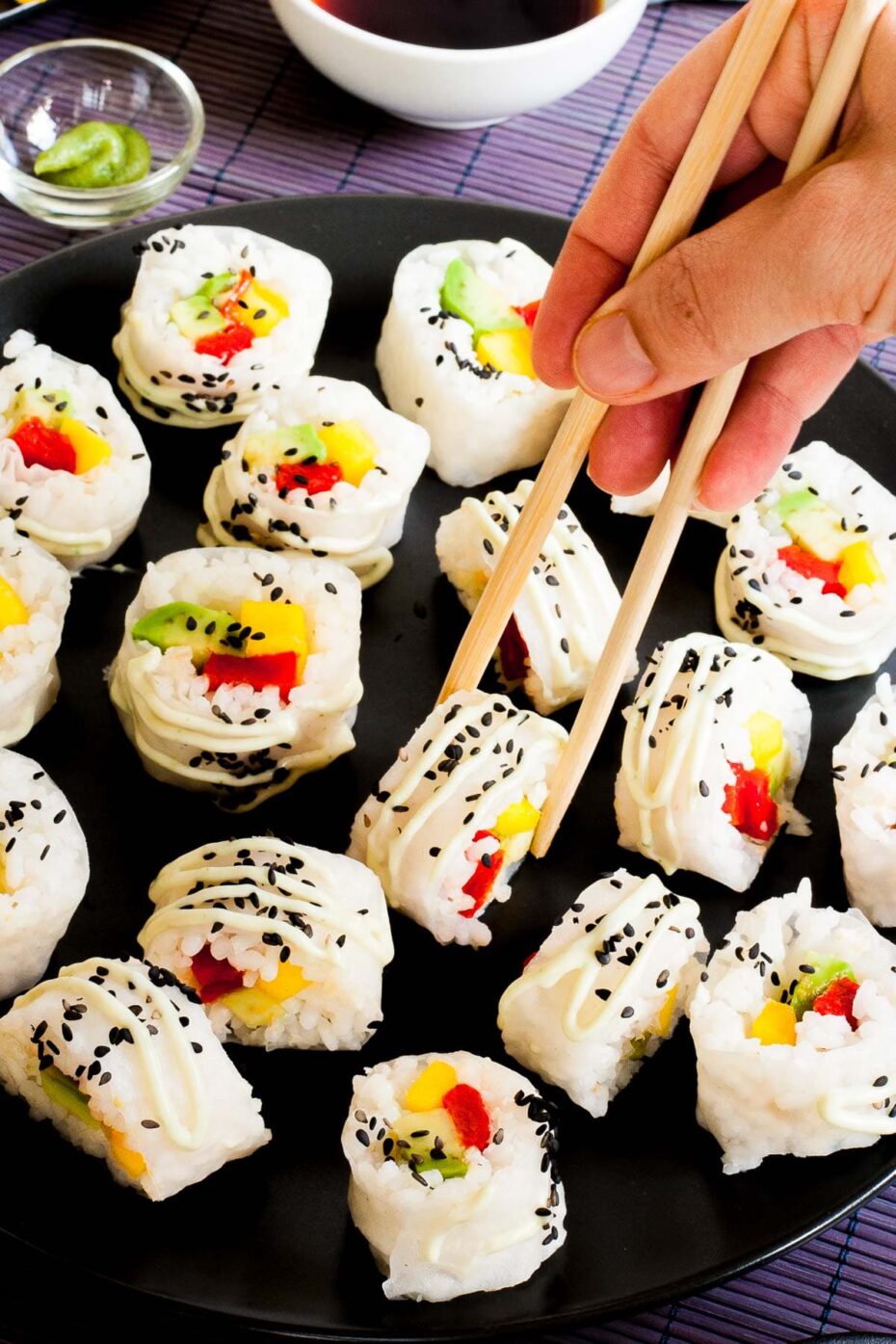 Large black round plate with a lot of white sushi rolls with colorful filling sprinkled with tiny black sesame seeds. A hand using chopsticks is taking one.