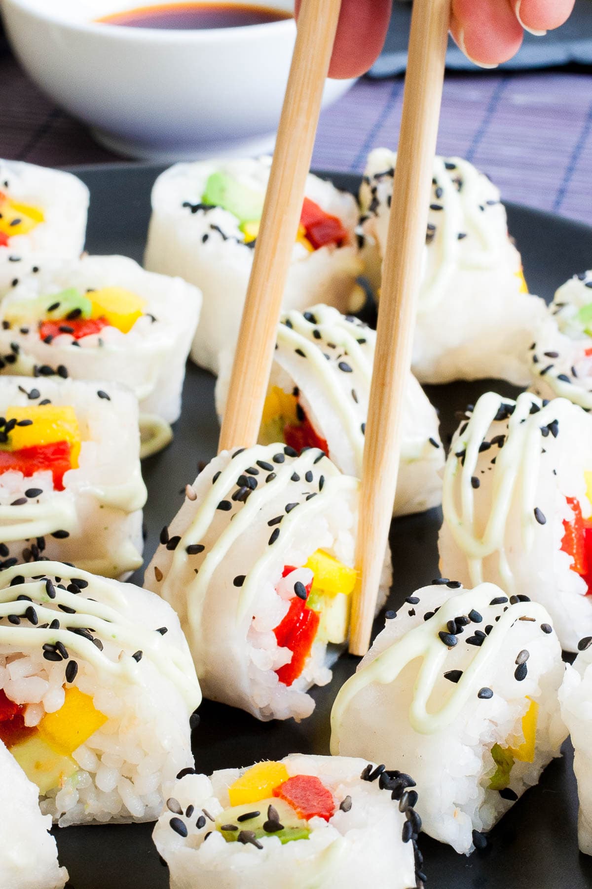 Large black round plate with a lot of white sushi rolls with colorful filling sprinkled with tiny black sesame seeds. A hand using chopsticks is taking one.