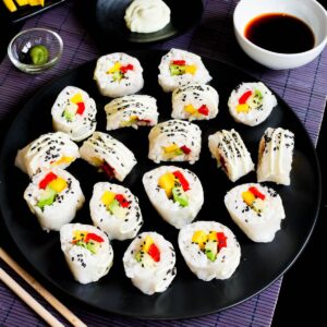 Large black round plate with a lot of white sushi rolls with colorful filling sprinkled with tiny black sesame seeds. Condiment bowls are around it.