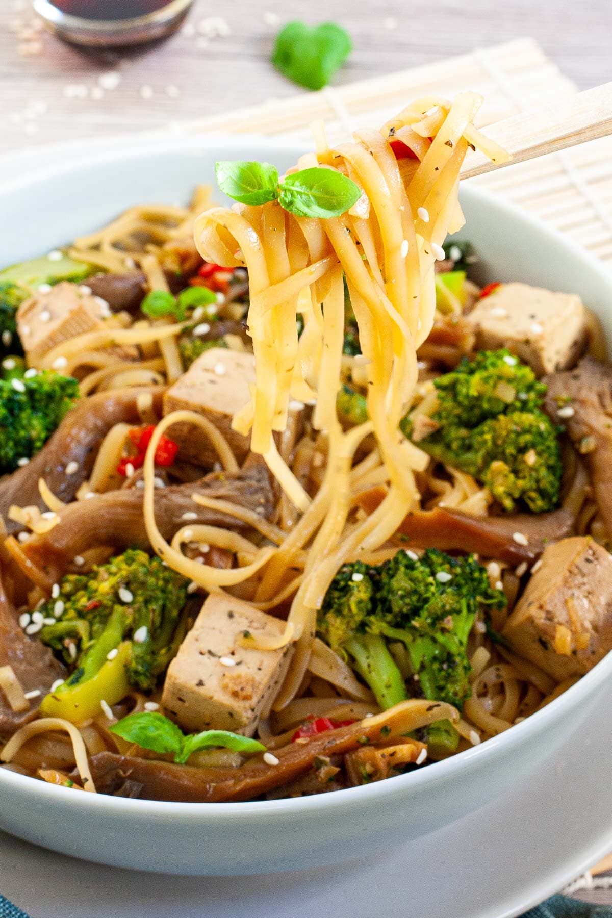 White bowl with tofu cubes, broccoli florets, oyster mushroom shreds, red chili slices. A wooden chopsticks are taking some of the rice noodles.