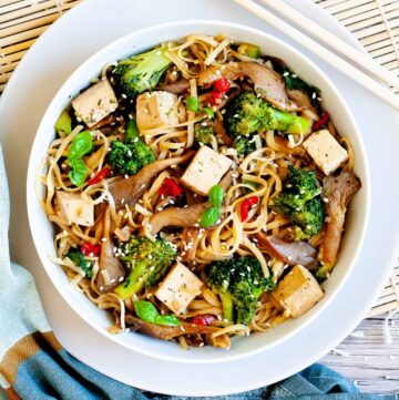 White bowl with tofu cubes, broccoli florets, oyster mushroom shreds, red chili slices and long thin rice noodles.
