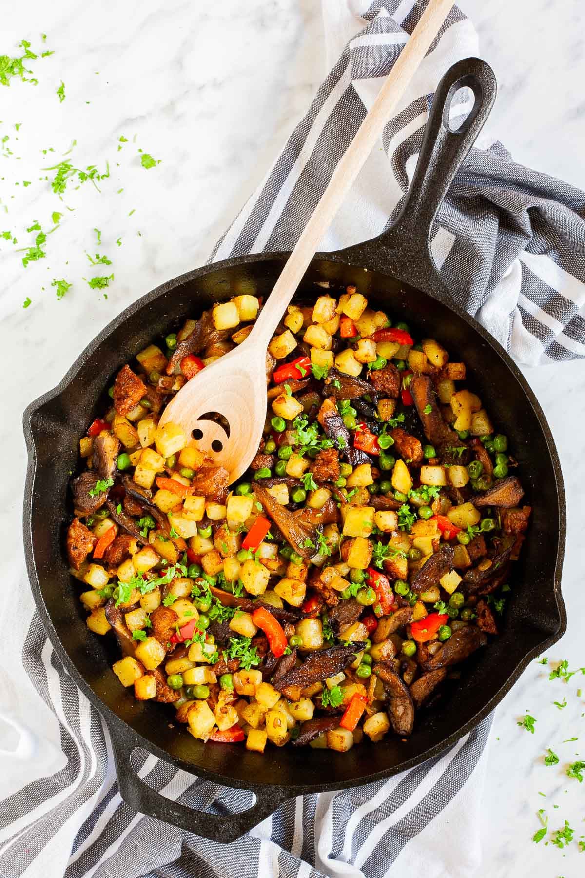 Black cast iron skillet from above with a wooden spatula and colorful chopped veggies like red bell pepper, yellow potatoes, brown mushroom, green peas, fresh parsley