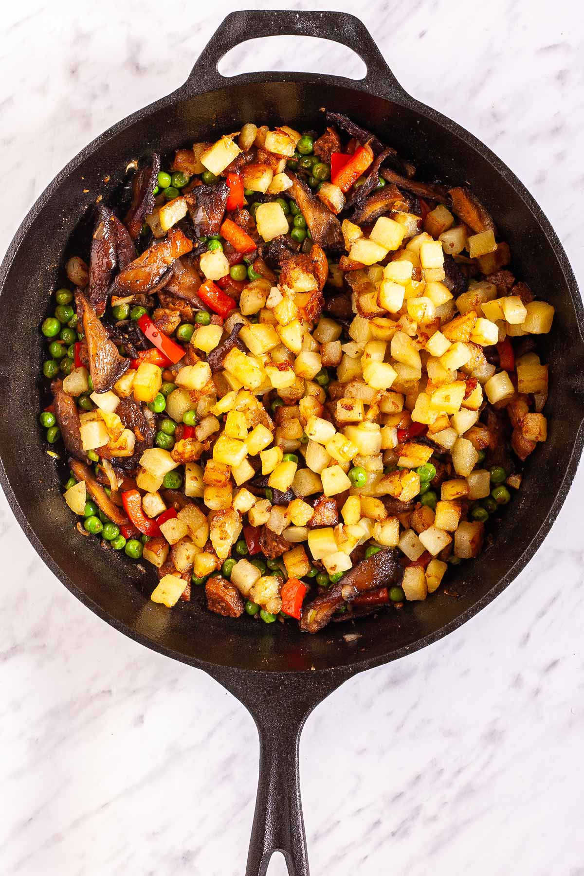 Black cast iron skillet with a mix of fried veggies like potatoes, green peas, chopped red pepper, chopped onion, crispy brown mushrooms and chopped vegan sausage.