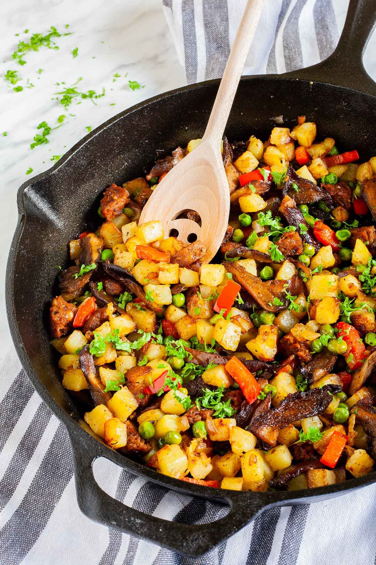 Black cast iron skillet from above with a wooden spatula and colorful chopped veggies like green peas, red bell pepper, yellow potatoes, brown mushroom, fresh parsley