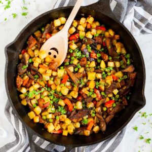 Black cast iron skillet from above with a wooden spatula and colorful chopped veggies like red bell pepper, yellow potatoes, brown mushroom, green peas, fresh parsley