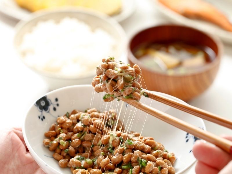 Sticky fermented soy beans are in a small white plate and a hand with chopsticks is taking some.