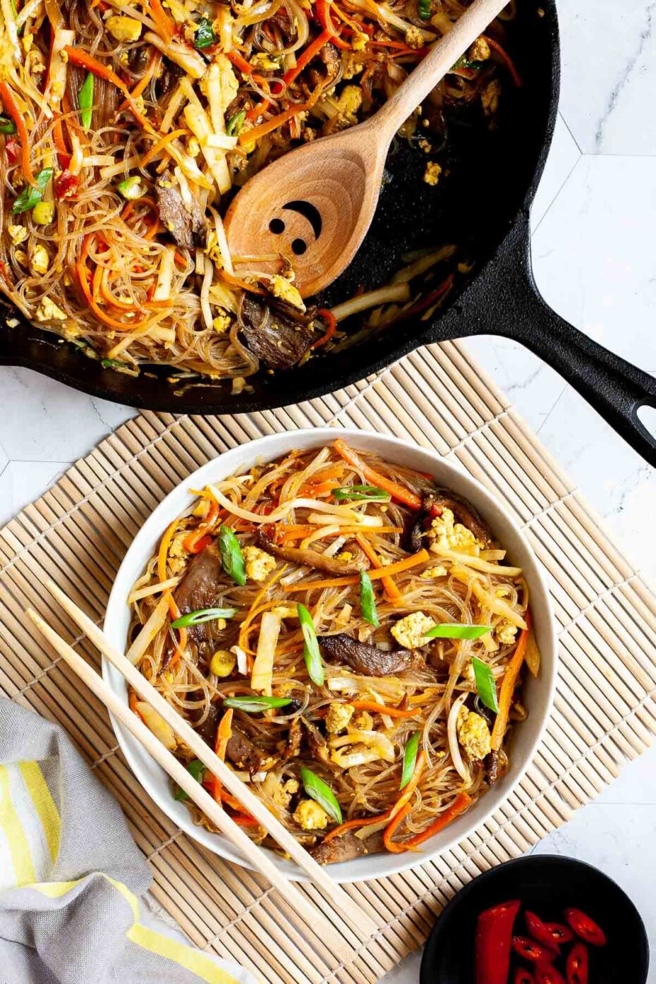 Black skillet from above full of glass noodles, shredded carrots, cabbage, sliced green onion, bean sprouts, mushroom shreds with a smiling wooden spatula. A white serving plate with chopsticks on it have the same contents