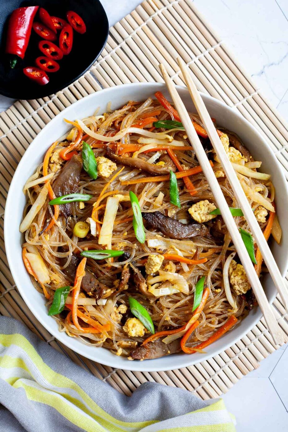 A white plate with chopstick from above is full of glass noodles, shredded carrots, cabbage, sliced green onion, bean sprouts, mushroom shreds.