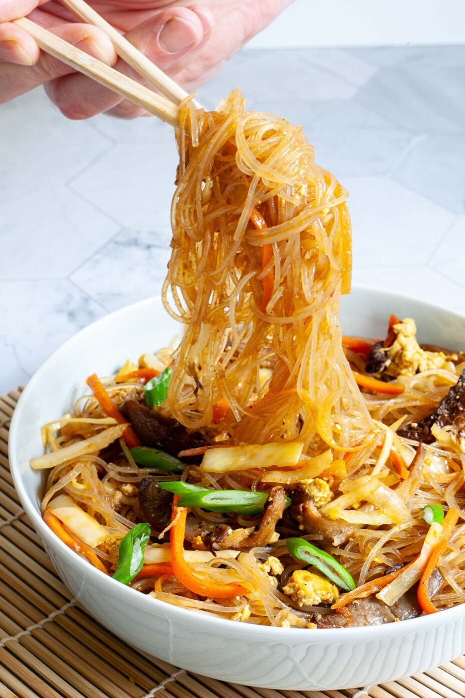 A white plate from above is full of glass noodles, shredded carrots, cabbage, sliced green onion, bean sprouts, mushroom shreds. A hand is holding chopsticks and lifting a generous amount of noodles up in the air.