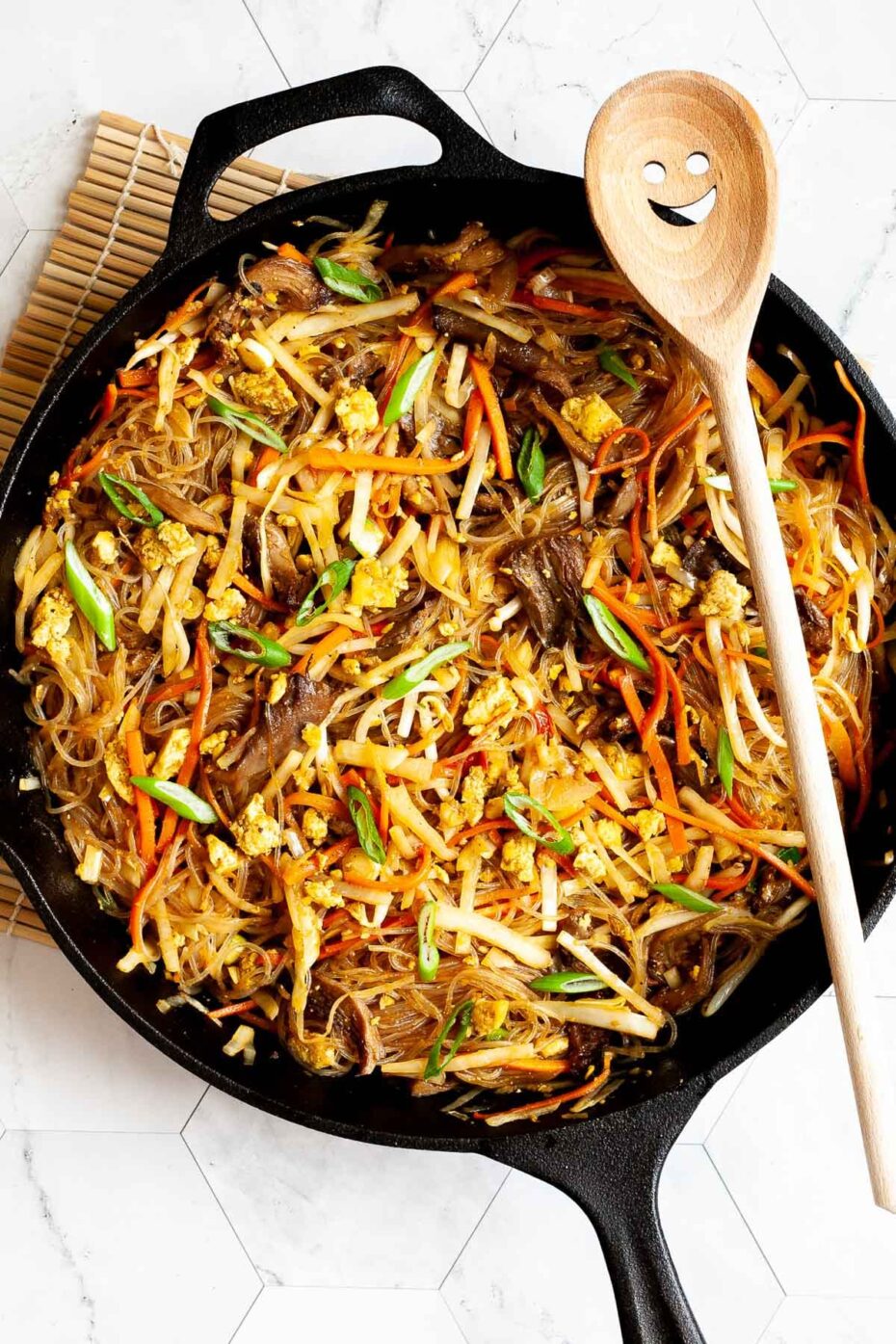 Black skillet from above full of glass noodles, shredded carrots, cabbage, sliced green onion, bean sprouts, mushroom shreds with a smiling wooden spatula