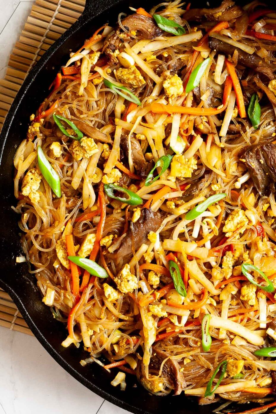 Black skillet from above full of glass noodles, shredded carrots, cabbage, sliced green onion, bean sprouts, mushroom shreds