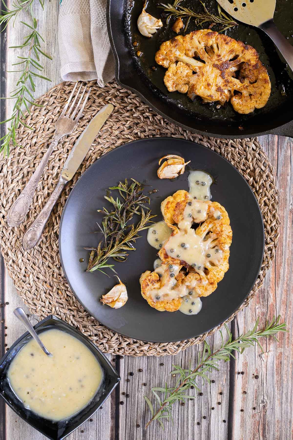 A crispy brown orange cauliflower slice is served on a black plate with roasted garlic cloves and wilted rosemary twigs. The leftover slices are in a cast iron skillet. A black bowl of green peppercorn sauce is place next to it.