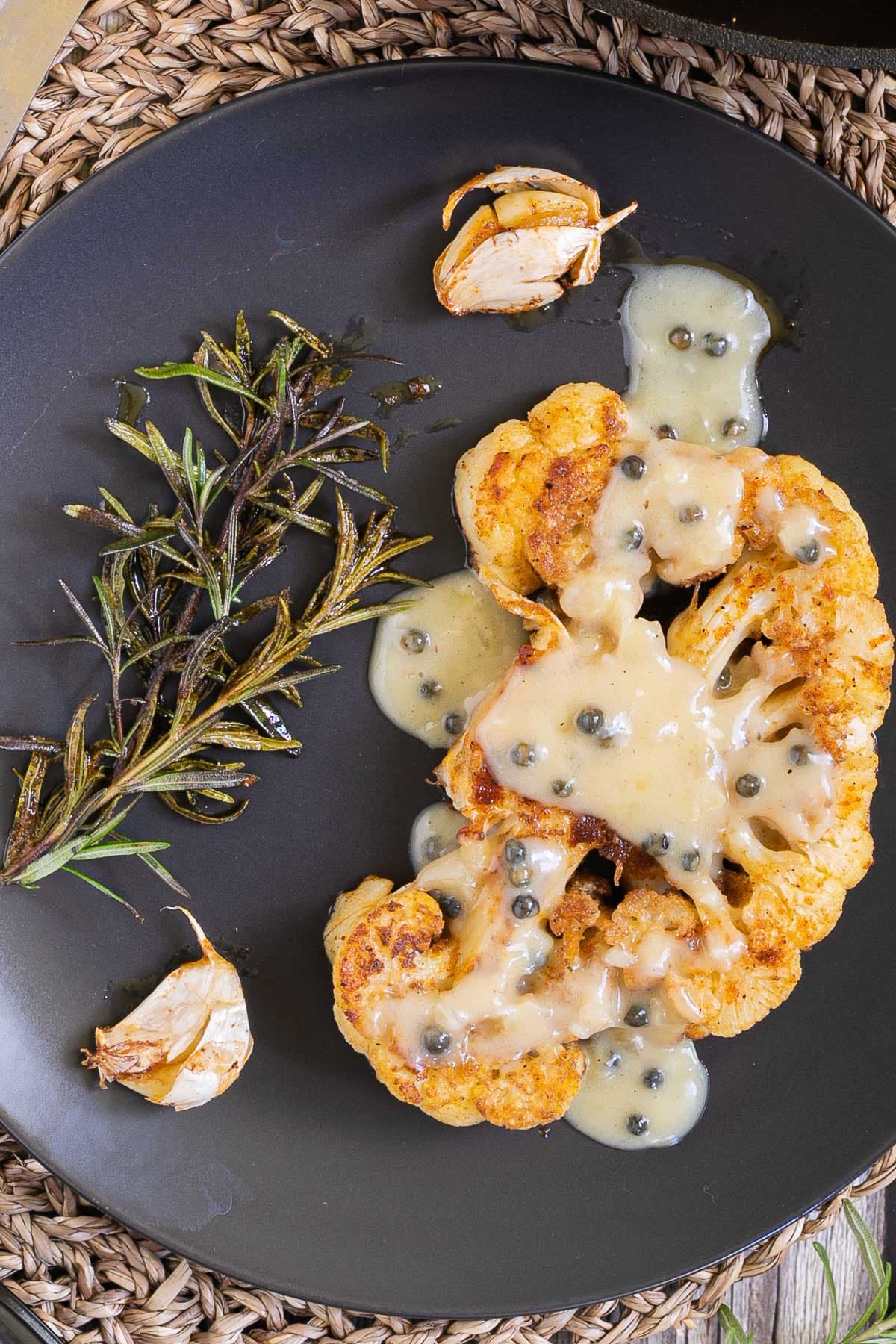 A crispy brown orange cauliflower slice is served on a black plate with roasted garlic cloves and wilted rosemary twigs.