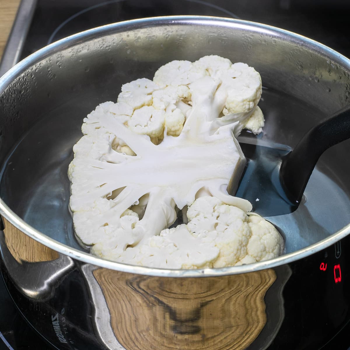 A slotted turner is submerging a large cauliflower slice in hot water.