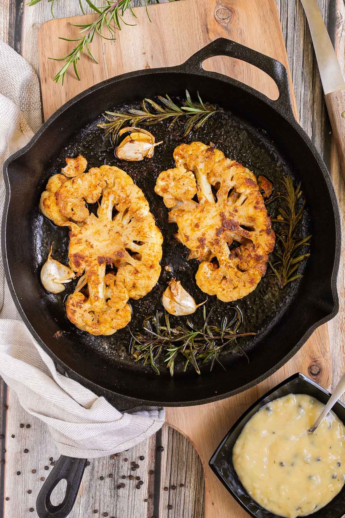 2 crispy brown yellow cauliflower slices in a cast-iron skillet surrounded by roasted garlic cloves and wilted rosemary twigs. A small black bowl of green peppercorn sauce is next to it.