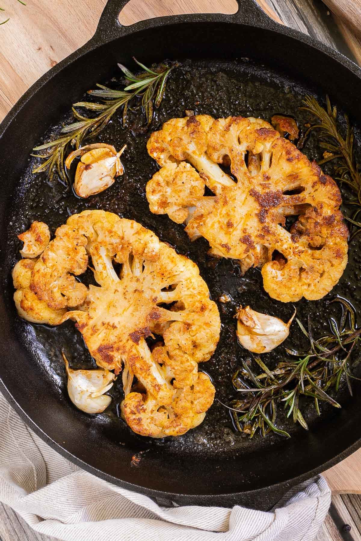 2 crispy brown yellow cauliflower slices in a cast-iron skillet surrounded by roasted garlic cloves and wilted rosemary twigs.
