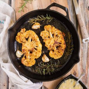 2 crispy brown yellow cauliflower slices in a cast-iron skillet surrounded by roasted garlic cloves and wilted rosemary twigs. A small black bowl of green peppercorn sauce is next to it.