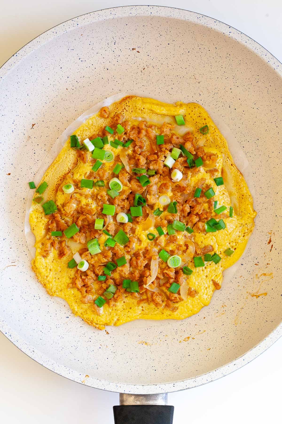 White frying pan with opaque rice paper topped with a yellow batter and sprinkled with brown crumbles and green spring onion rings