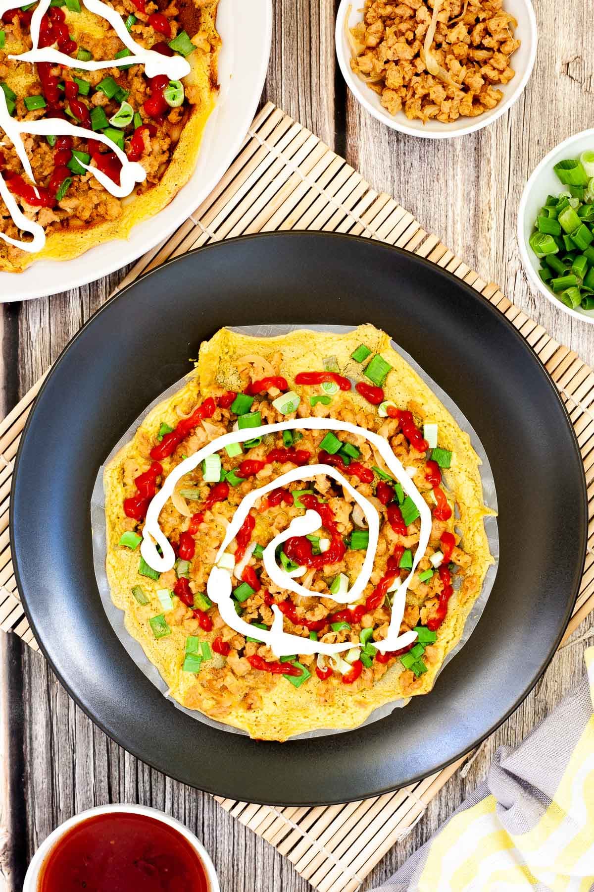 Black plate with opaque rice paper topped with a yellow batter and sprinkled with brown crumbles and green spring onion rings. White and red sauces are drizzled on top. 